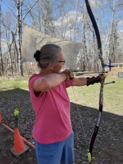 Sweetgrass Fitness Archery Women with pink shirt ready to shoot a bow and arrow at an outdoor archery range.