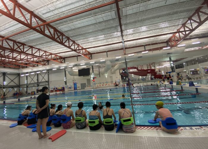 Women-in-Motion-Picture Swim Against the Stream 2023 Backs of women in swimming suits sitting on the edge of a pool deck listening to a swim instructor who is in the water.