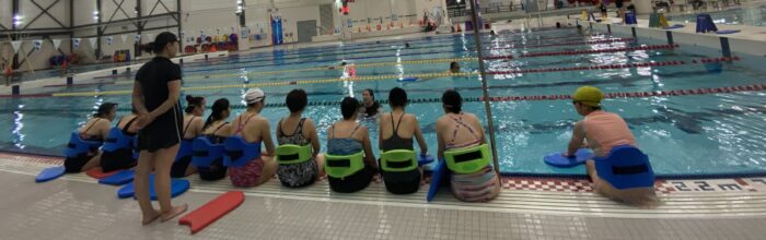Backs of women in swimming suits sitting on the edge of a pool deck listening to a swim instructor who is in the water.