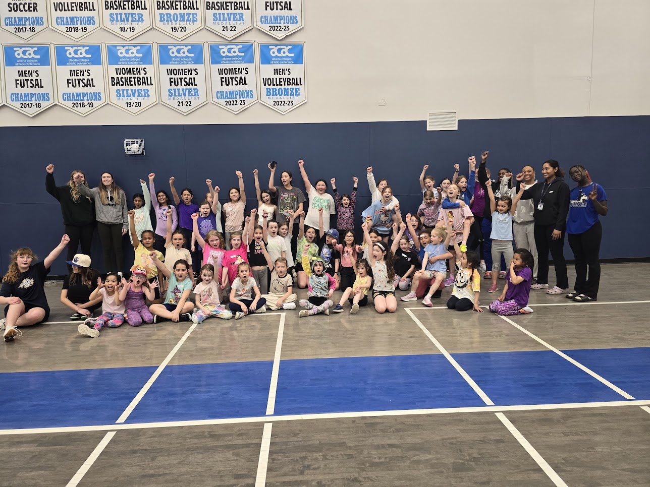 Group of 50 girls in a gym posing for a picture in athletic wear and their arms raised with a fist pump