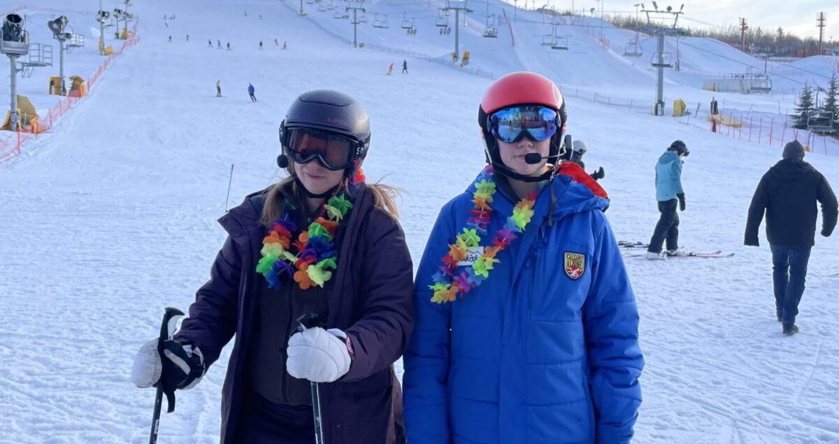 Two women at the base of a ski hill in ski gear wearing rainbow leis around their neck.