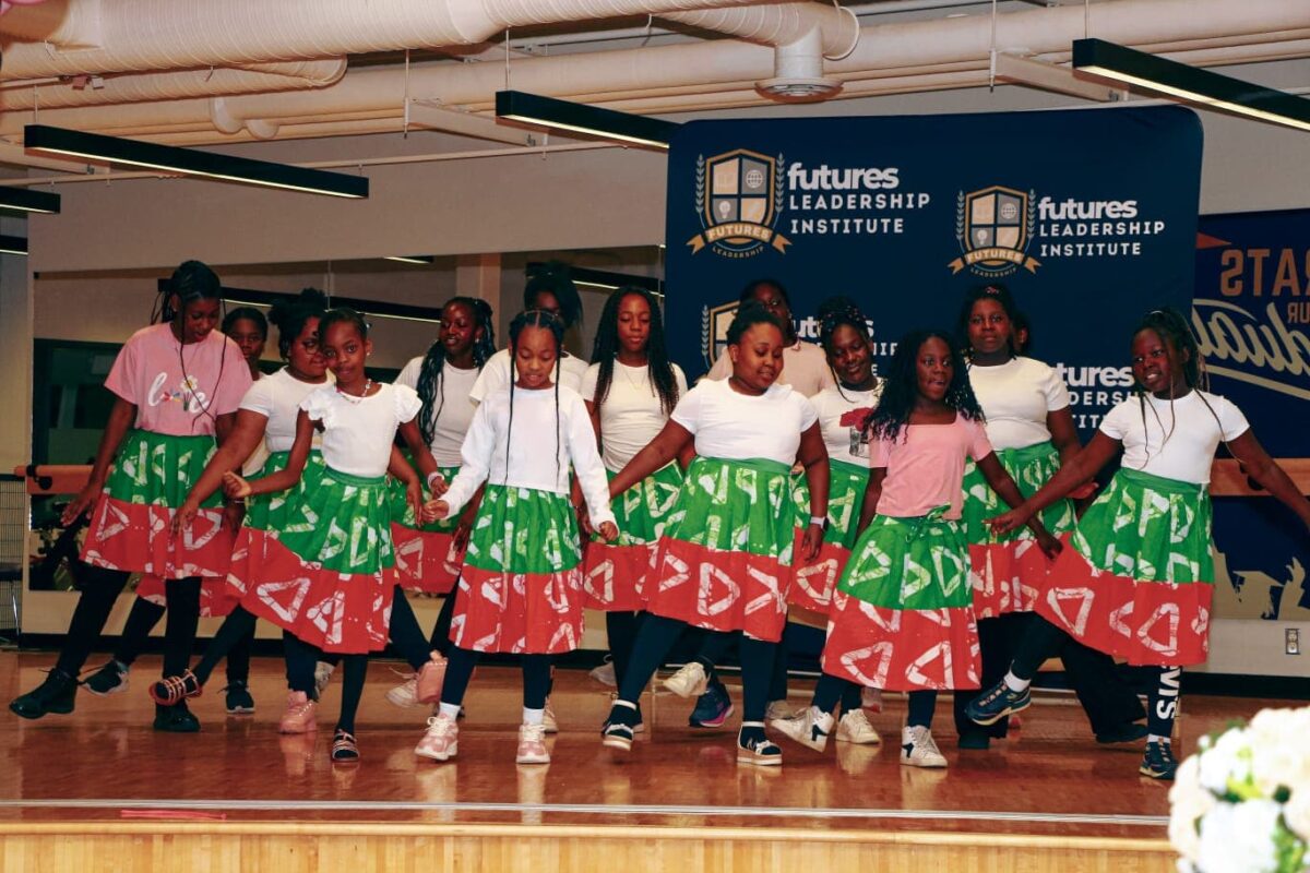 Group of young Black girls in pink and green skirts performing a dance on stage.