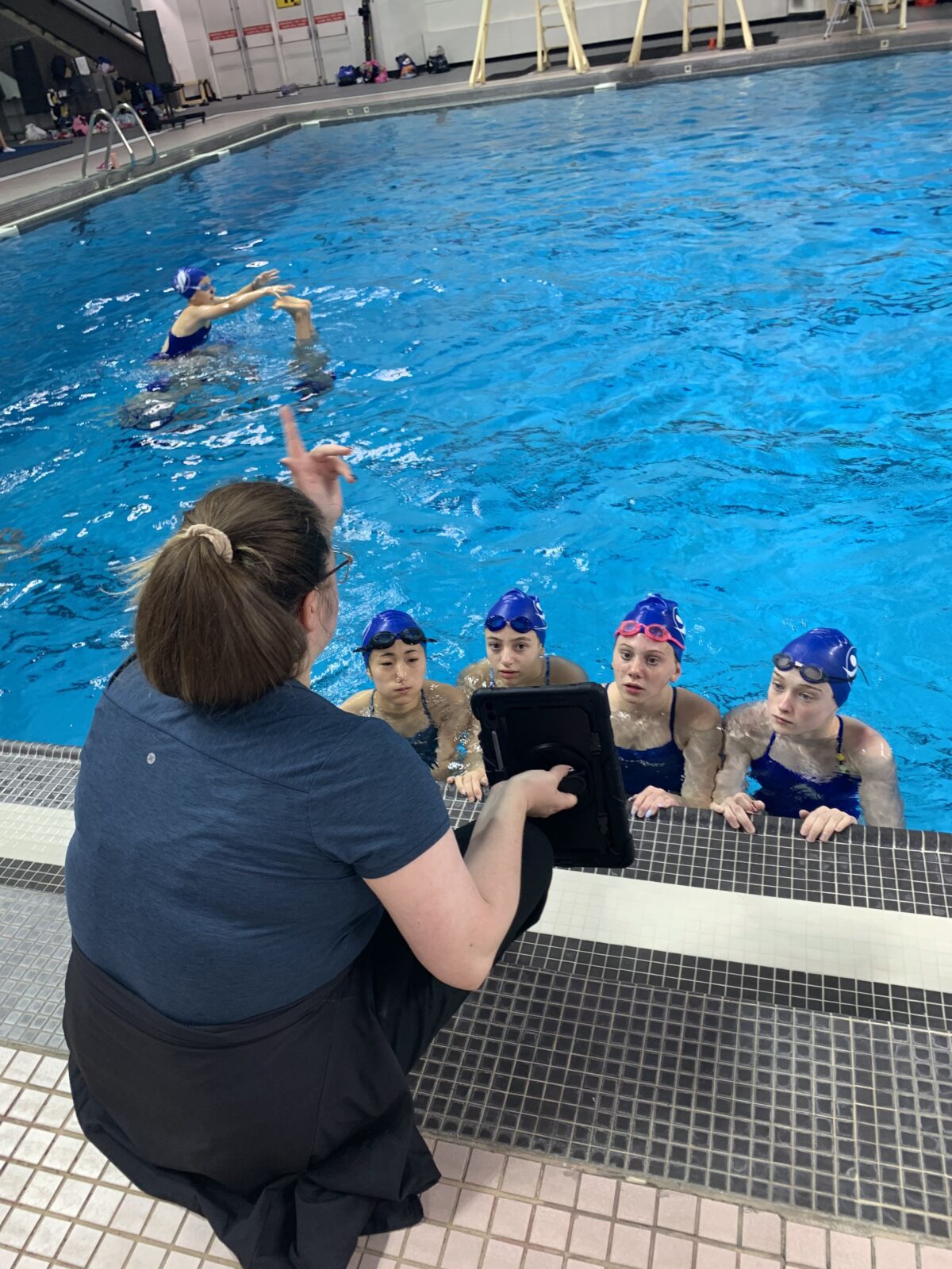 Artistic Swimming coach bending down with an ipad showing a group of 3 girls in the pool the screen.