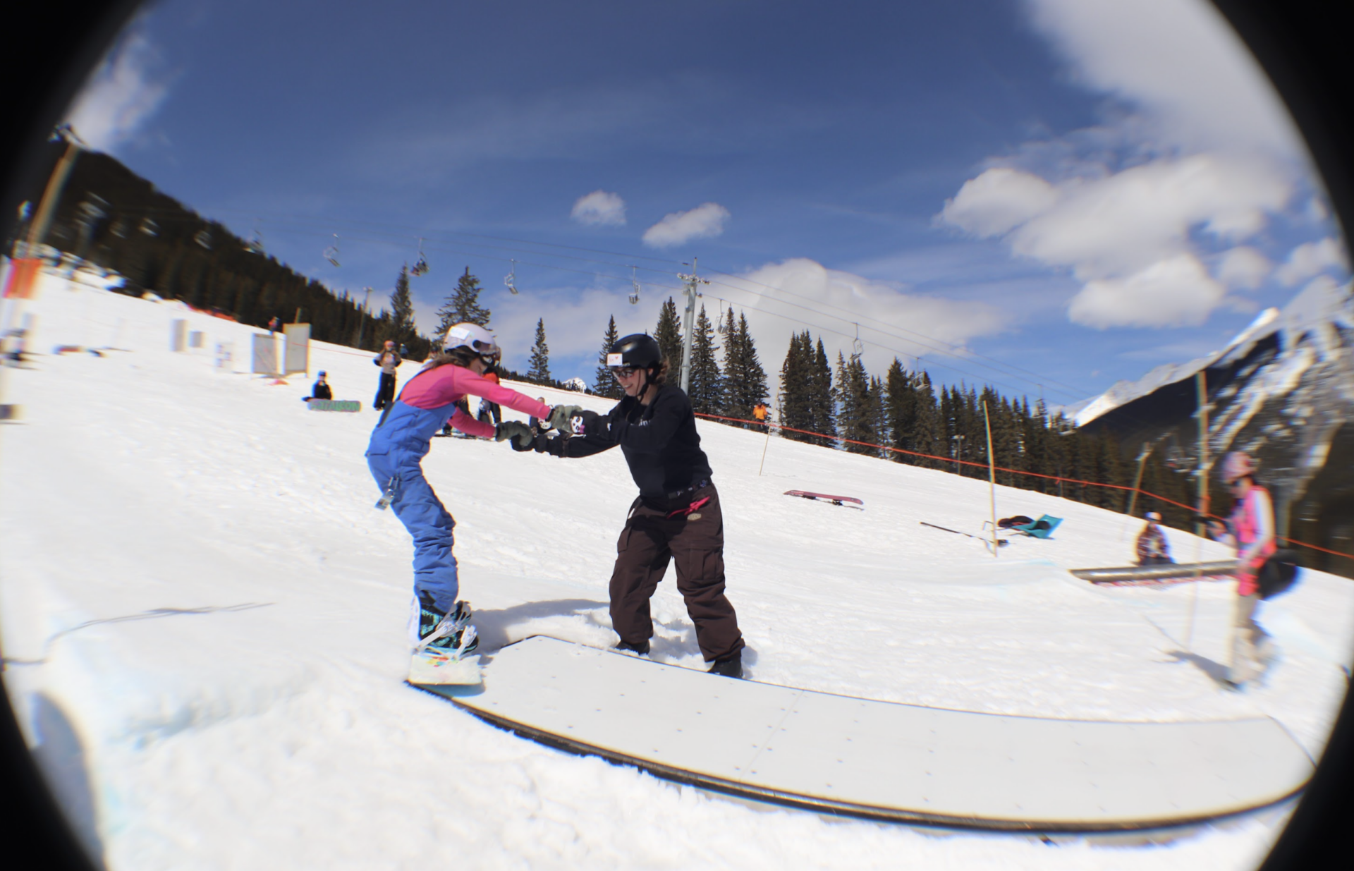 Youth girl helping a younger girl learn to snowboard.