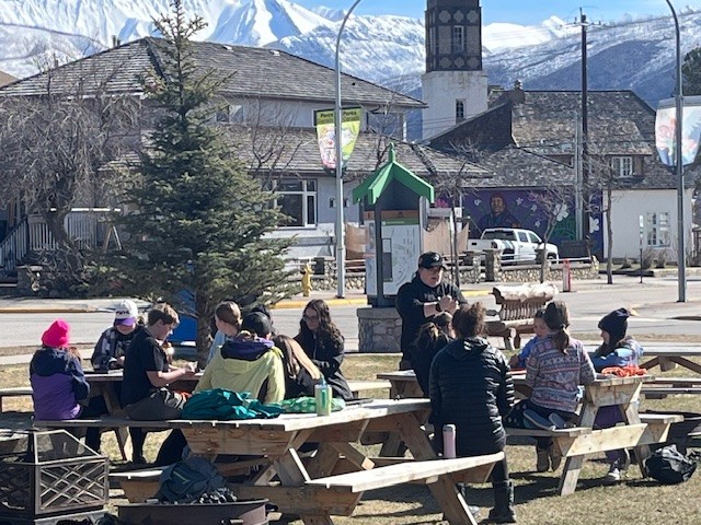 Girls outside listening to an adult speak while sitting at picnic tables, with mountains in the background. 